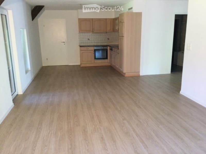 Empty room with wooden flooring, white walls, and a small kitchen area with cabinets and appliances visible in the background.