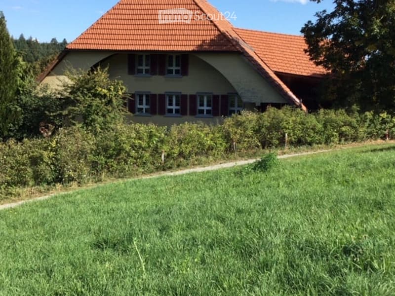 A two-story house with a red-tiled roof and beige walls, surrounded by greenery and a grassy lawn.