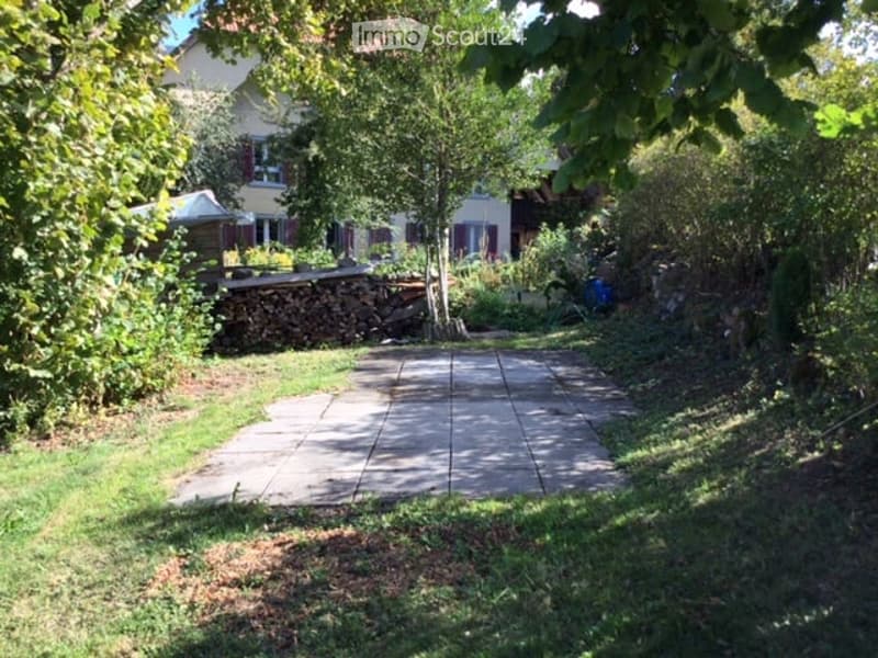 Garden area with a paved pathway leading to a house, surrounded by greenery, trees, and a stack of firewood.