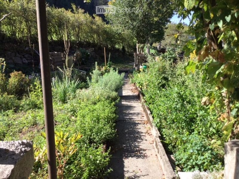 A well-maintained garden with a gravel pathway flanked by lush greenery, including shrubs, herbs, and small trees. The garden is enclosed by stone walls and wooden fences, with a serene, natural setting.
