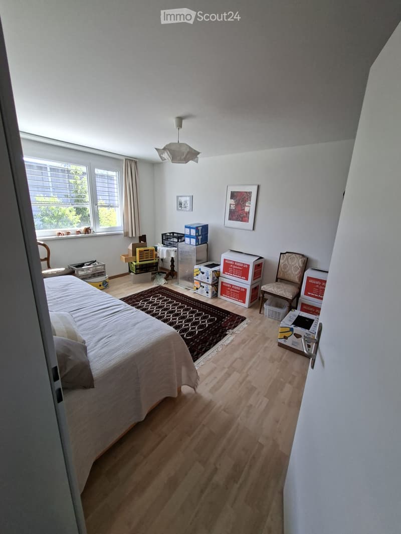 Bedroom with wooden flooring, a bed with pillows and a blanket, a chair, a small table with a lamp, and several boxes stacked near the window. There is a large window with blinds and a curtain, and a picture frame on the wall.
