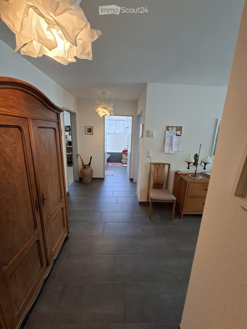 Hallway with tiled floor, wooden wardrobe, small wooden chair, table lamp, and a view into a bedroom with a bed and bathroom.