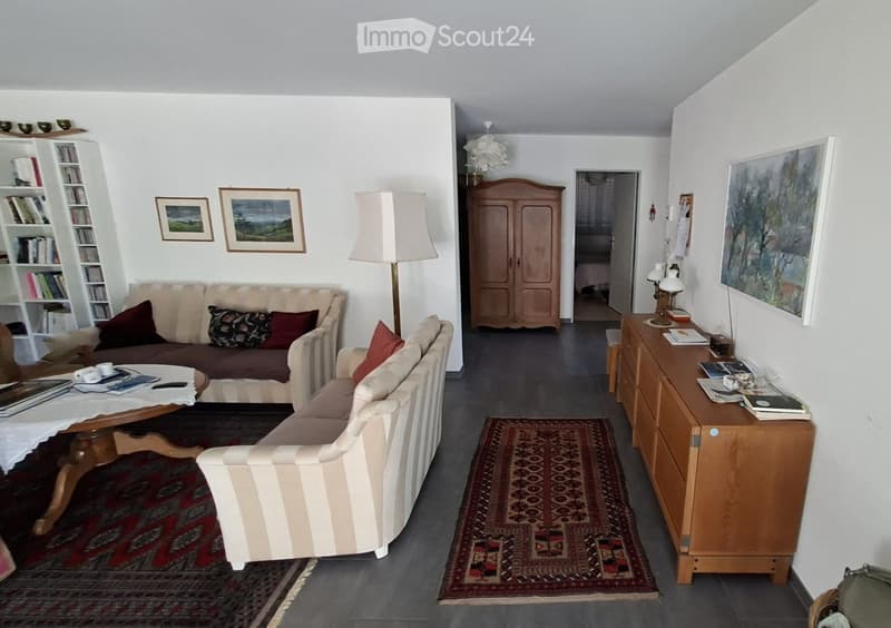 Living room with a sofa, coffee table, armchair, cabinet, bookshelf, and lamps. The room features a patterned rug and several picture frames on the walls.