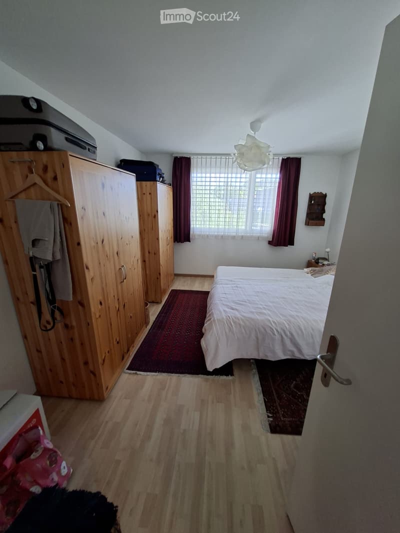 Cozy bedroom with wooden flooring, a large bed with white bedding, a wooden wardrobe, a window with dark curtains, and a small nightstand with a lamp.