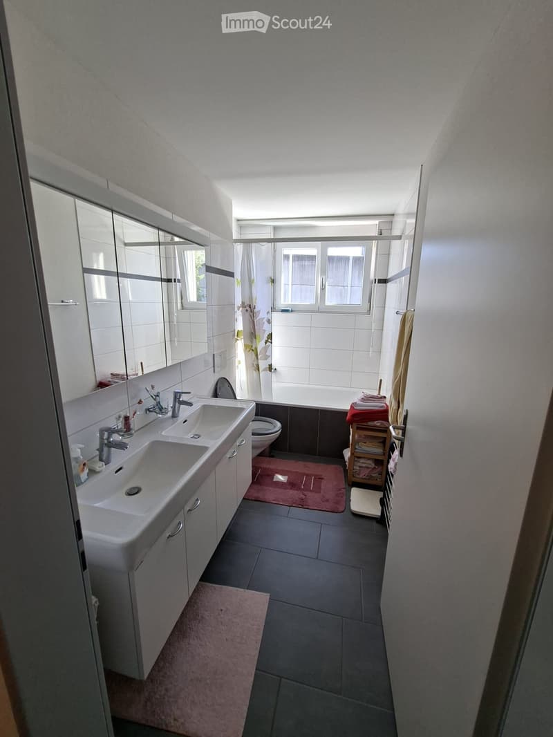 Modern bathroom with double sink, mirror, toilet, bathtub, and towels neatly arranged. Features include a large window and a small shelf with folded towels.