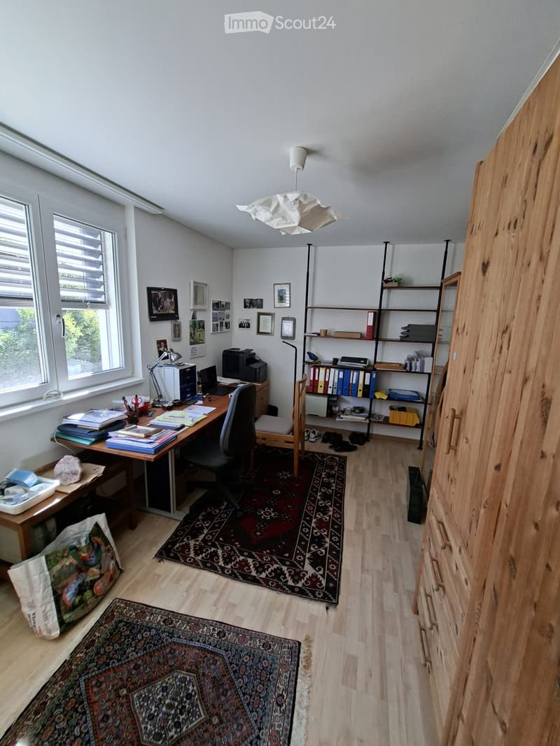 Well-lit office space with wooden flooring, large windows, a desk with a laptop, printer, and books, a comfortable chair, shelves filled with binders and books, and decorative rugs.