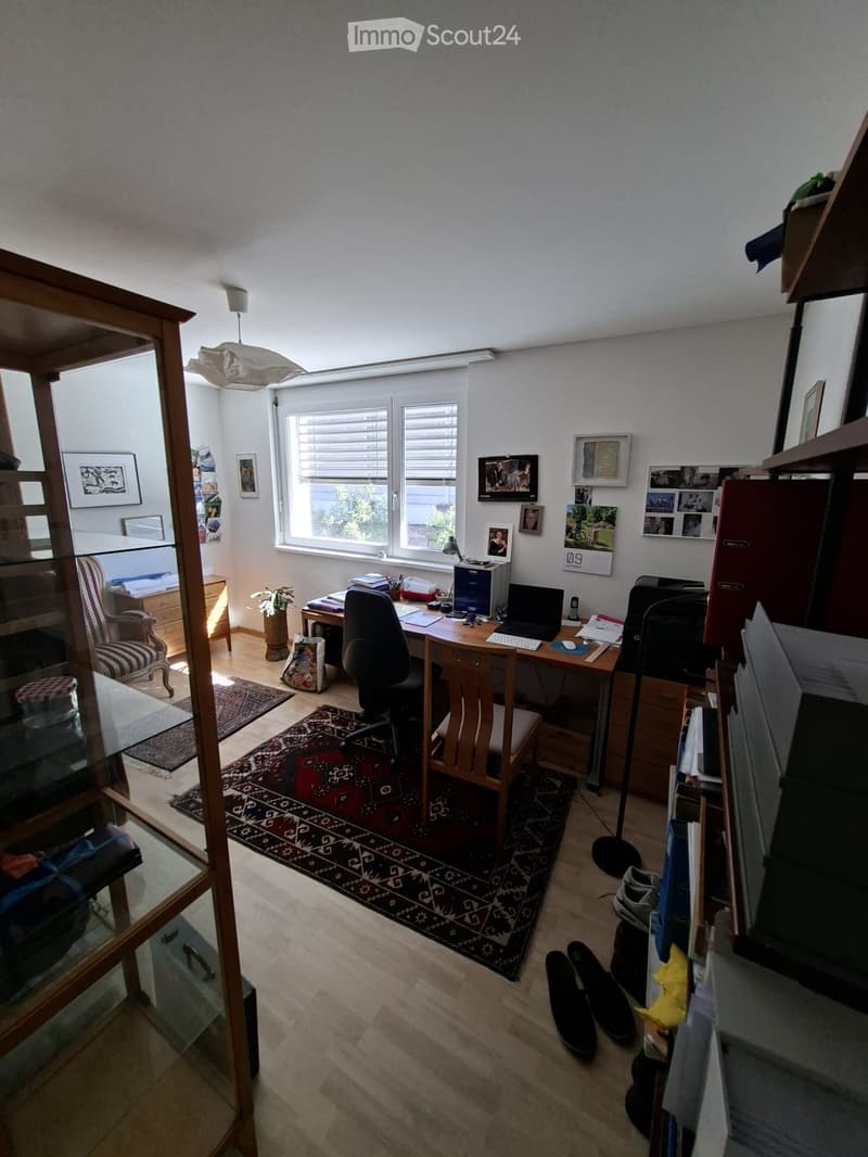 A well-furnished room with a wooden desk, office chair, laptop, printer, bookshelf, and various decorative items including framed pictures and a calendar. The room features wooden flooring, a large window with blinds, and a patterned rug.