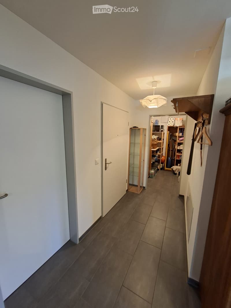 A narrow hallway with tiled flooring, white walls, and several doors leading to different rooms. A small glass display cabinet and a coat rack with hangers are visible on the right side.