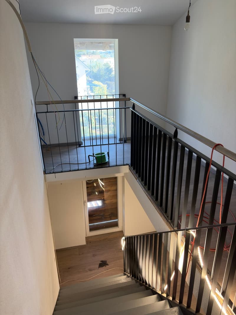 Modern staircase with wooden steps and metal railing, leading to a balcony area with a view of the outside. The walls are painted white, and the lighting is bright.