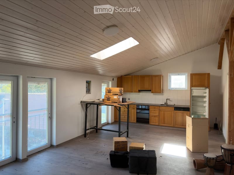 The room features a wooden ceiling, white walls, and a large window. It includes a table with boxes, a kitchen area with cabinets, an oven, a refrigerator, and a sink.