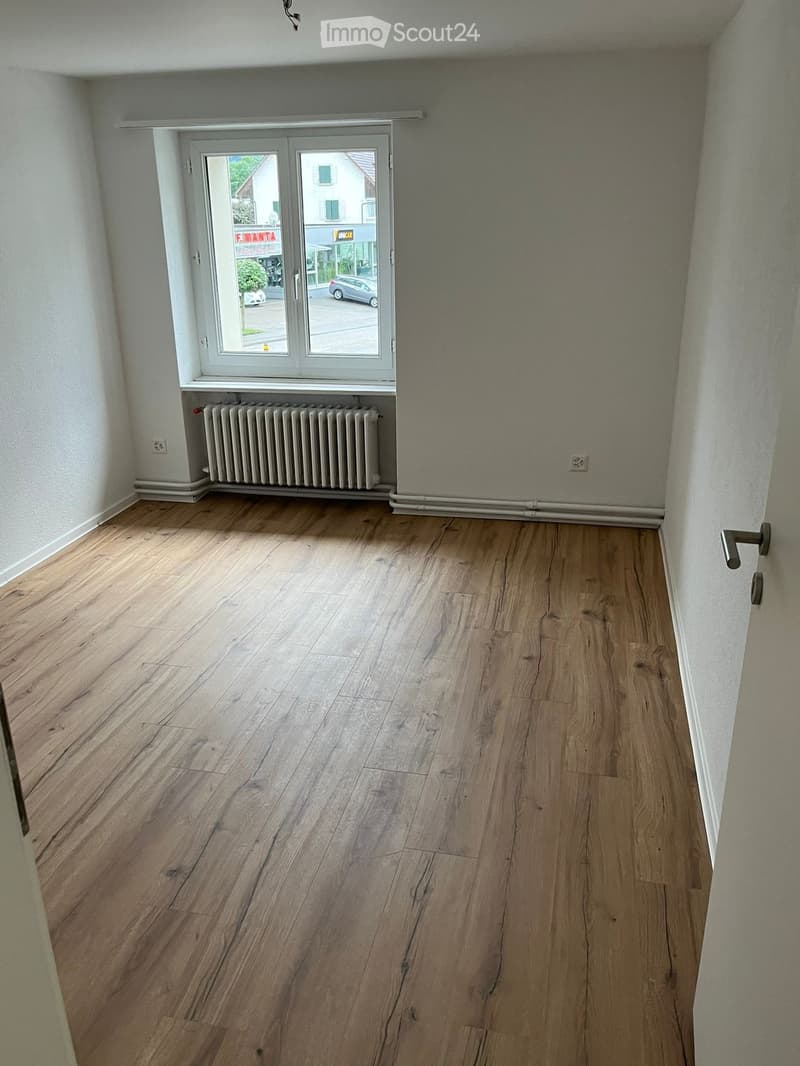 Empty room with wooden flooring, white walls, a radiator, and a large window with a view of the street.