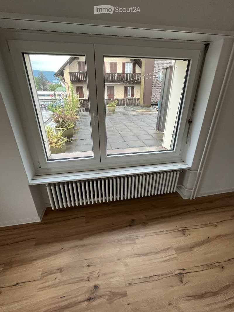 Empty room with wooden flooring, radiator, and large windows offering a view of the outdoor area and surrounding buildings.