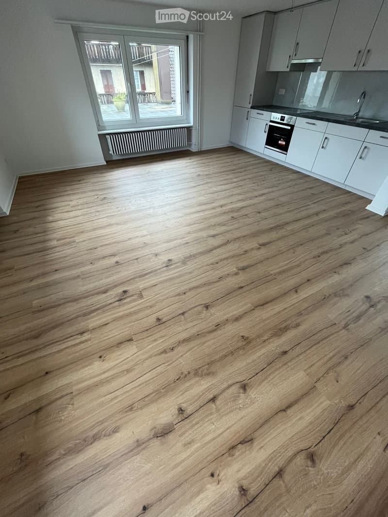 Empty kitchen with light wooden flooring, white cabinets, and a window with a view of the balcony.