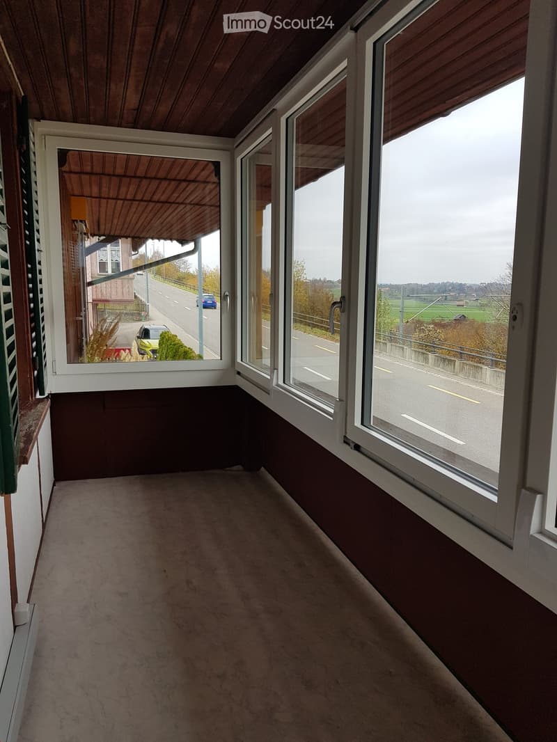 Balcony with large windows offering views of a road and surrounding landscape, carpeted floor, wooden ceiling.