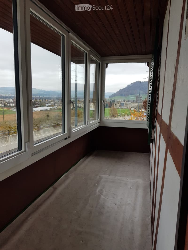 Empty balcony with large windows offering a scenic view of a town and surrounding landscape.