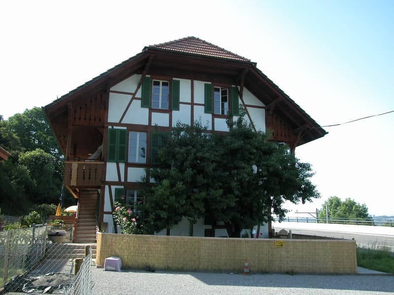 Two-story traditional house with white and brown timber framing, green shutters, and a wooden balcony accessible via stairs.