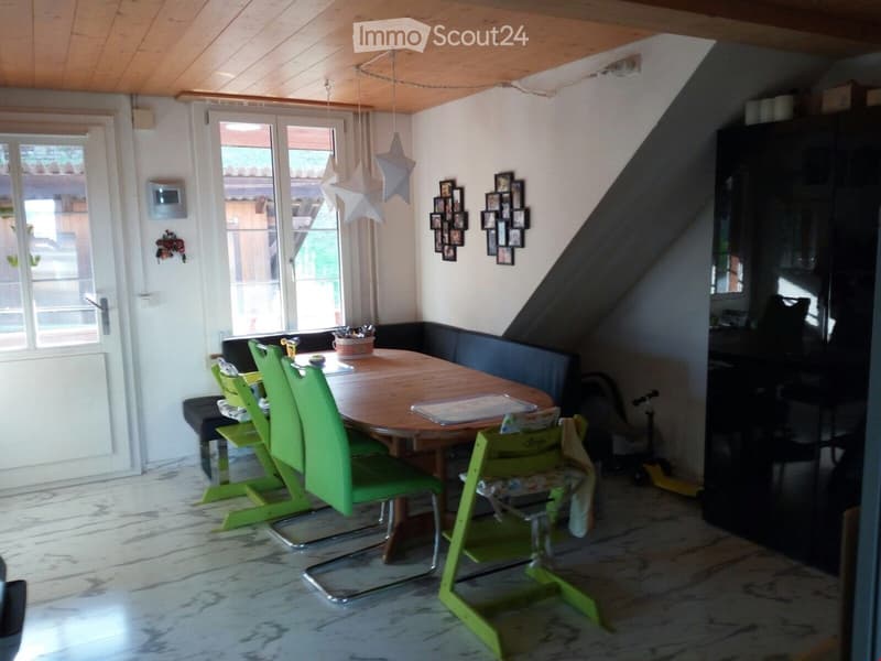 Brightly lit dining area with a round wooden table surrounded by green chairs, a black bench seating, and a small black cabinet. The room features a white tiled floor, a wooden ceiling, and large windows with a view of the outdoor area.