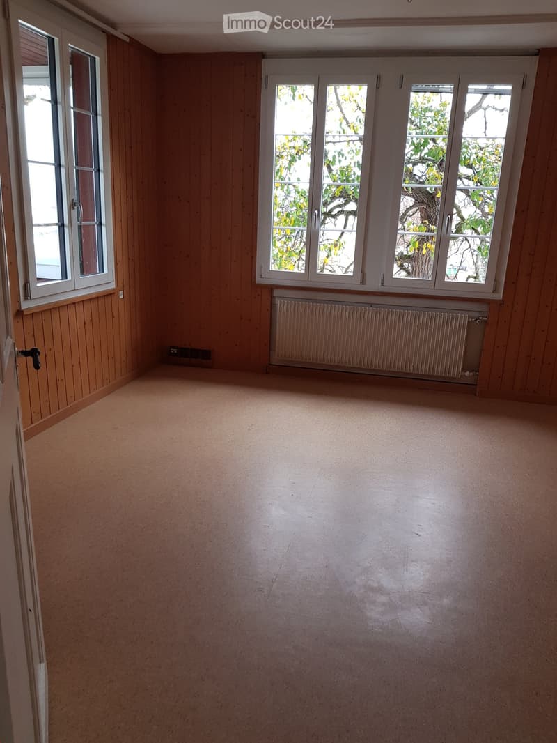 Empty room with wooden panel walls, carpeted floor, large windows with white frames, radiator, and a door with a black handle.