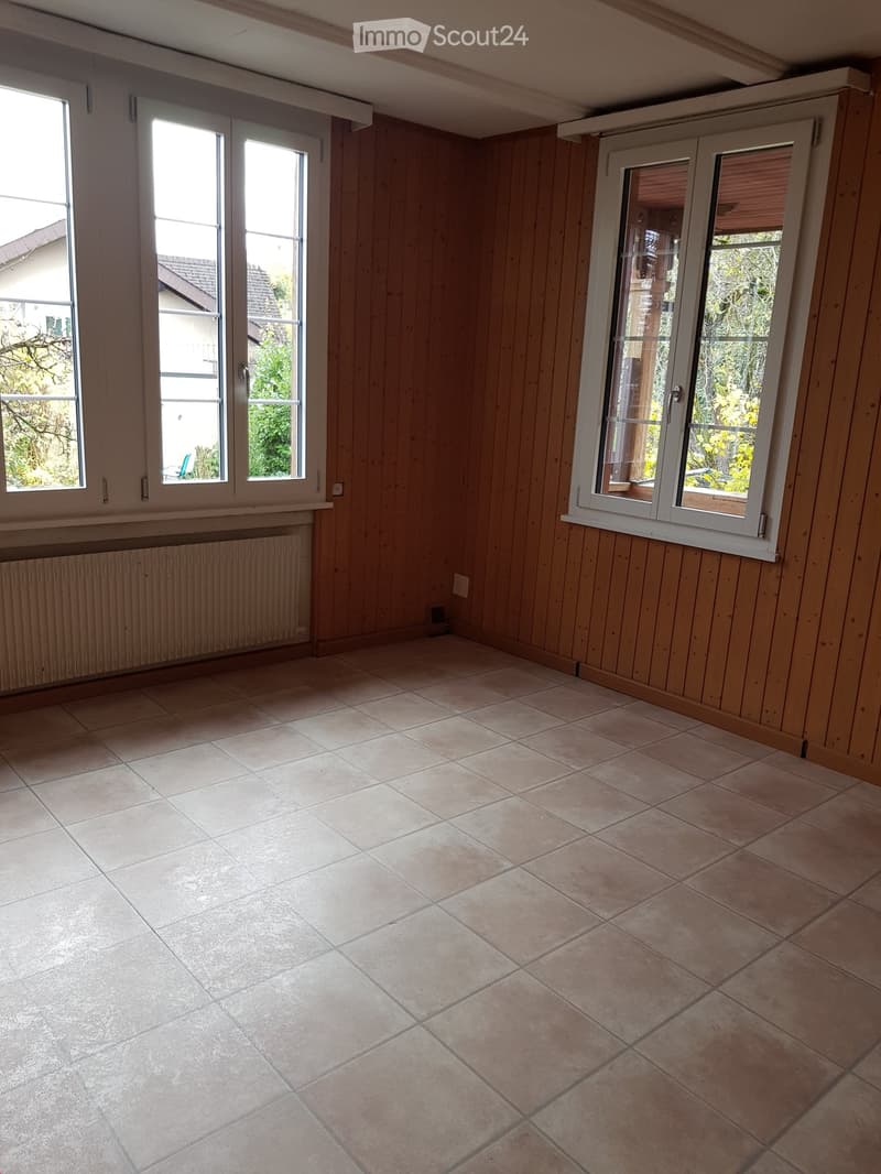 Empty room with wooden panel walls, tiled floor, and large windows with white frames. Natural light illuminates the space.