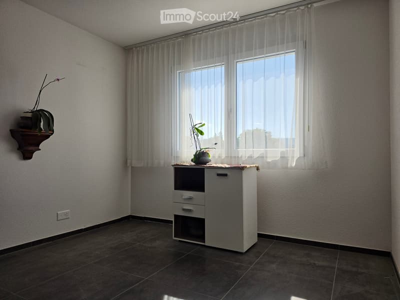 A small, unfurnished room with a tiled floor, a window with sheer curtains, a small cabinet with drawers, and a shelf holding a potted plant.