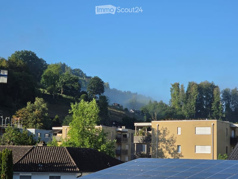A multi-story residential building with balconies and solar panels on the roof, surrounded by trees and greenery.