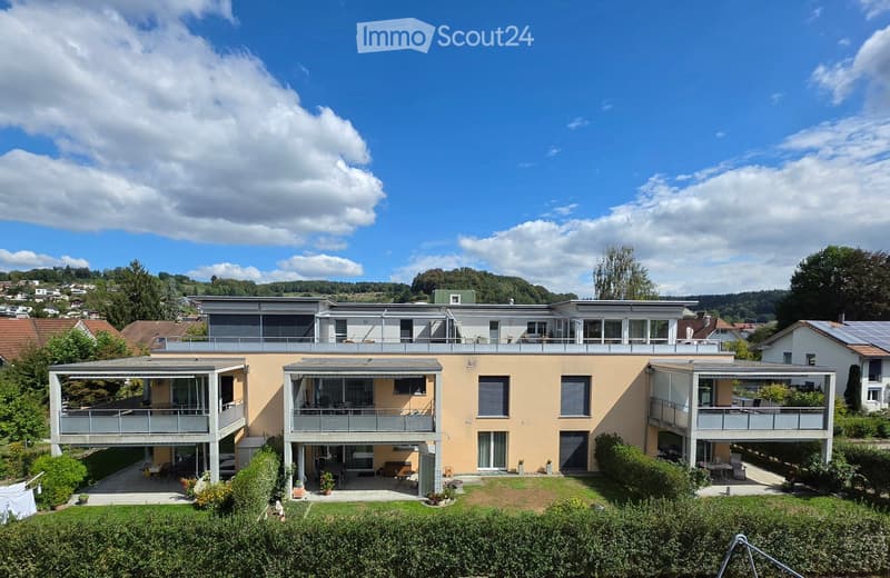 Modern two-story house with beige facade, multiple balconies, large windows, and surrounding greenery.
