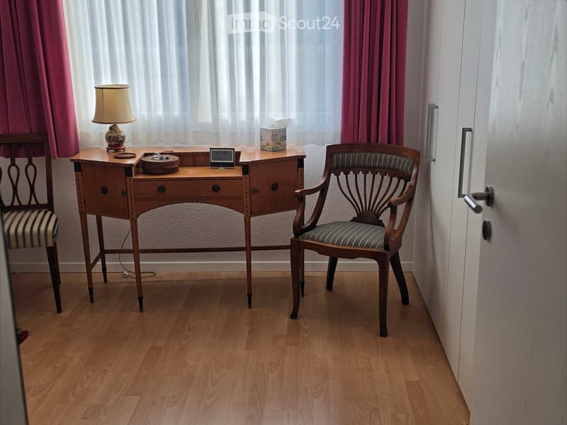 A small, well-lit room with wooden flooring, a wooden desk, two chairs, a lamp, and a small clock on the desk.