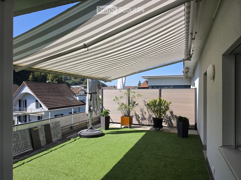 Balcony with artificial grass, covered by a retractable awning, featuring potted plants and an umbrella stand.