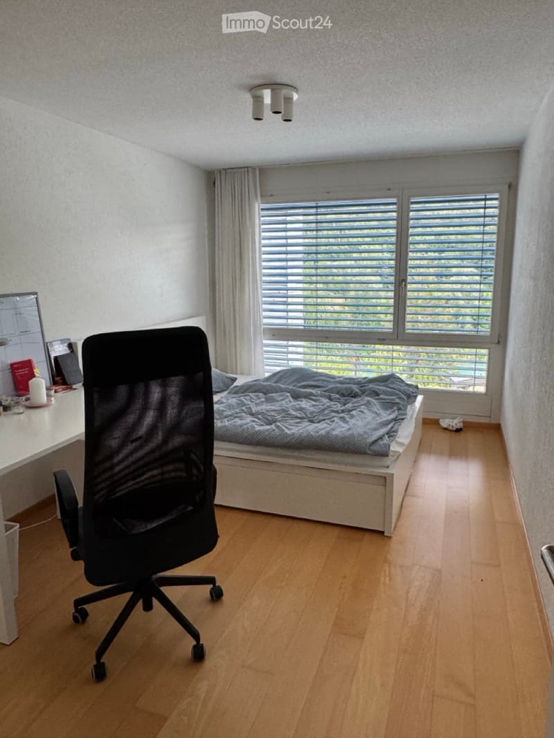 A modern bedroom with a wooden floor, a bed with a grey duvet, a desk with a screen, a black office chair, and a window with blinds and curtains.