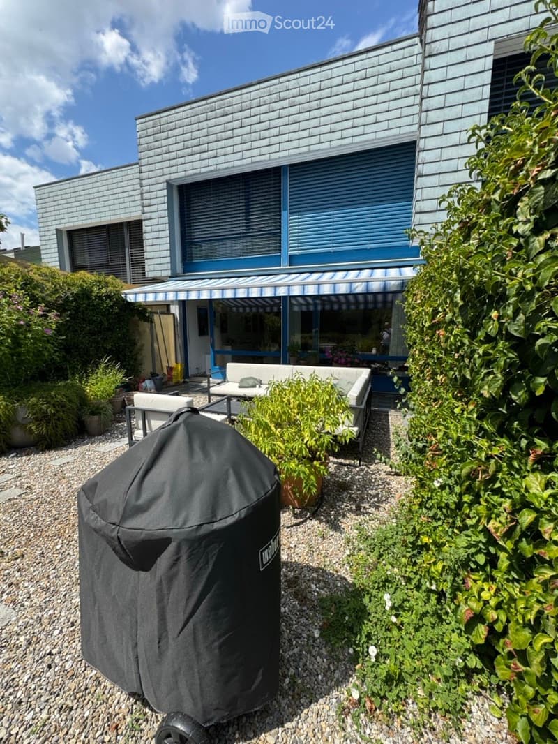 Outdoor patio area with a covered seating area, featuring a sofa, table, chairs, and a covered grill, surrounded by plants and a modern house with blue shutters.