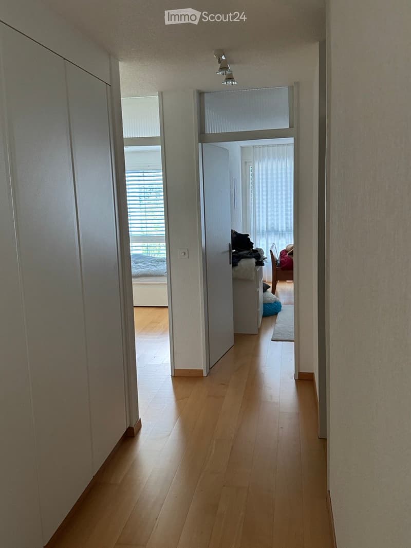 A narrow hallway with wooden flooring, white walls, and a ceiling light fixture. The hallway leads to a partially visible bedroom with a bed and a small sitting area with a chair and a small table.