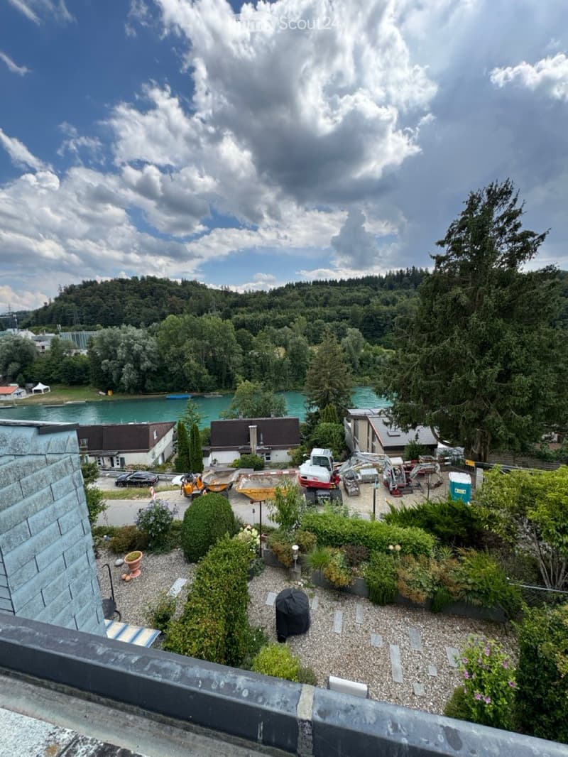 A rooftop garden with gravel ground, potted plants, and shrubs, overlooking a lake and surrounding greenery.