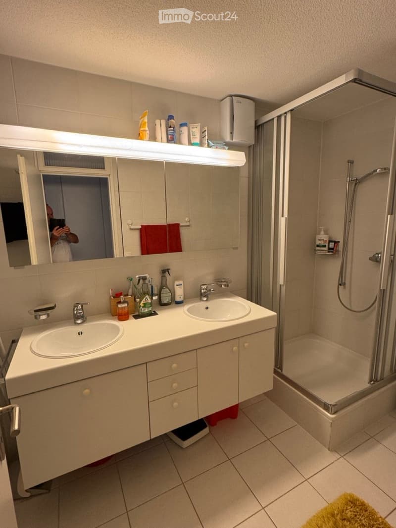 Modern bathroom with double sink, large mirror, shower, and various toiletries on the counter.