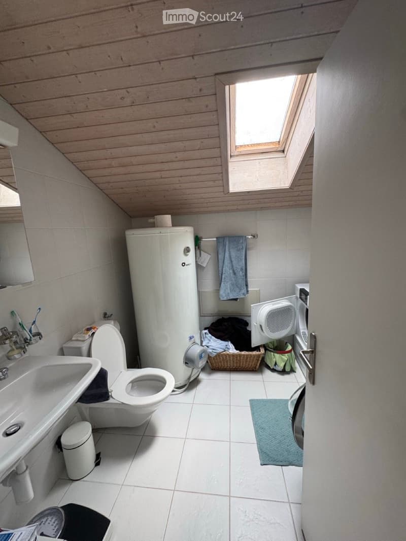 Bathroom with white tiled floor and walls, wooden ceiling, skylight window, sink, toilet, mirror, washing machine, wicker basket, and towel.