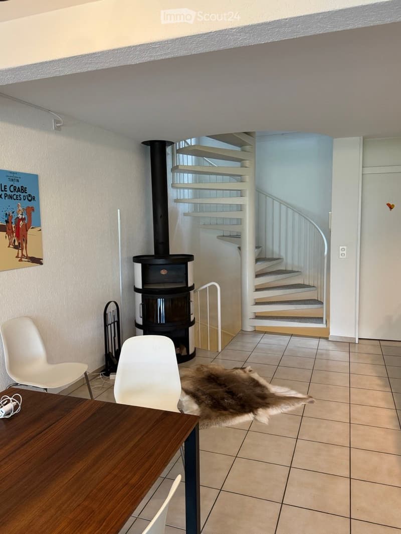 A modern, well-lit room with a tiled floor, a wooden table, two white chairs, a fireplace, a fur rug, and a spiral staircase leading upwards.