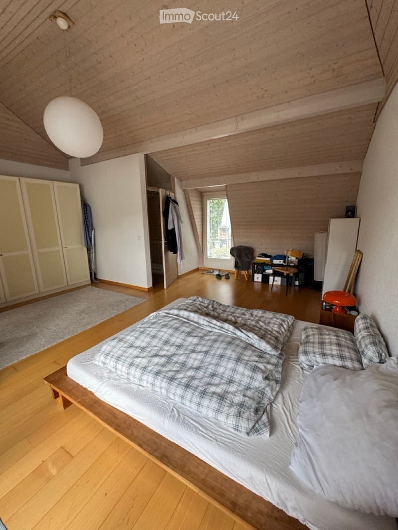 Cozy attic bedroom with wooden floors, slanted wooden ceiling, large window, bed with plaid bedding, wardrobe, small table, and chair.