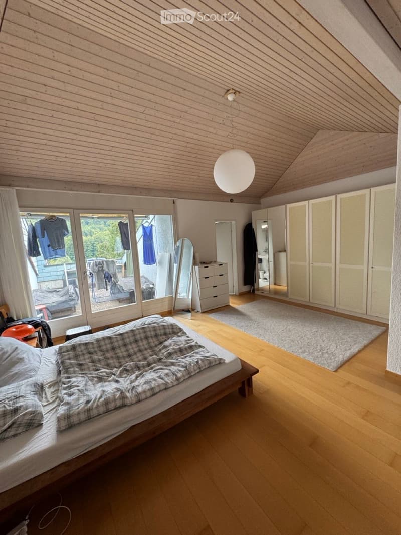 Bright bedroom with wooden floors, slanted wooden ceiling, large window with clothes hanging, bed with checkered blanket, wardrobe, dresser, and round pendant light.