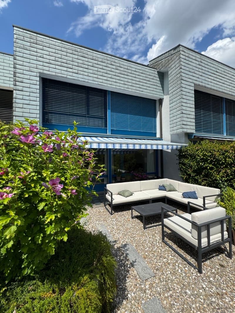 Modern outdoor patio with a grey sectional sofa, coffee table, and chairs, surrounded by greenery and a striped awning attached to a contemporary house.