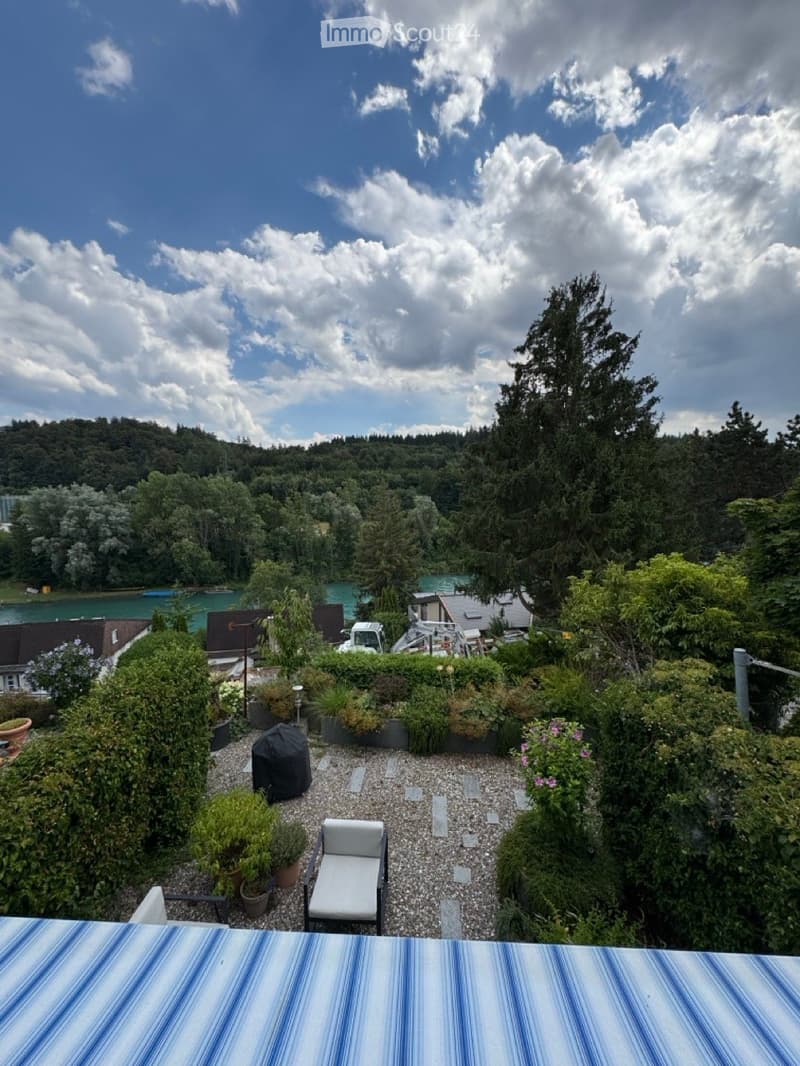 A serene garden area with gravel pathways, potted plants, a lounge chair, and a scenic view of a lake and forested hills under a partly cloudy sky.