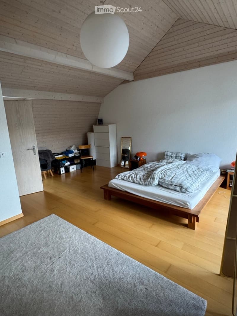 Cozy attic bedroom with wooden floors, slanted wooden ceiling, and a large round pendant light. Features a bed with a patterned duvet, a wooden chair, a small table with a lamp, a mirror, and a cabinet.