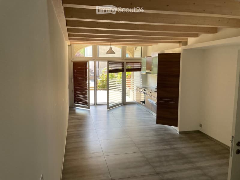 Bright hallway with tiled flooring, wooden ceiling beams, and large glass doors leading to an outdoor area. The hallway connects to a modern kitchen with stainless steel appliances.