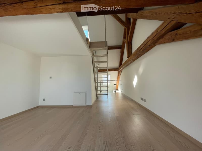 Empty room with wooden flooring, white walls, exposed wooden beams, and a metal staircase leading to an upper level.