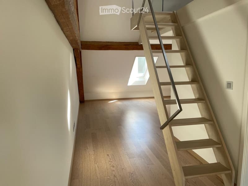 Spacious hallway with wooden flooring, exposed wooden beams, and a modern staircase with a metal railing leading upwards. Natural light streams in from a skylight at the top of the stairs.