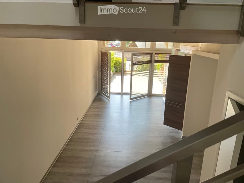 Bright hallway with tiled floor, white walls, and large glass doors leading to an outdoor area. Wooden railing visible on the staircase.