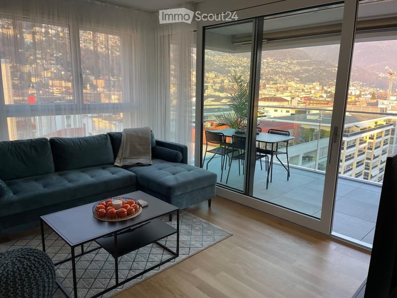 Modern living room with a grey sectional sofa, coffee table with fruit bowl, and a small outdoor dining set on a balcony overlooking a cityscape.