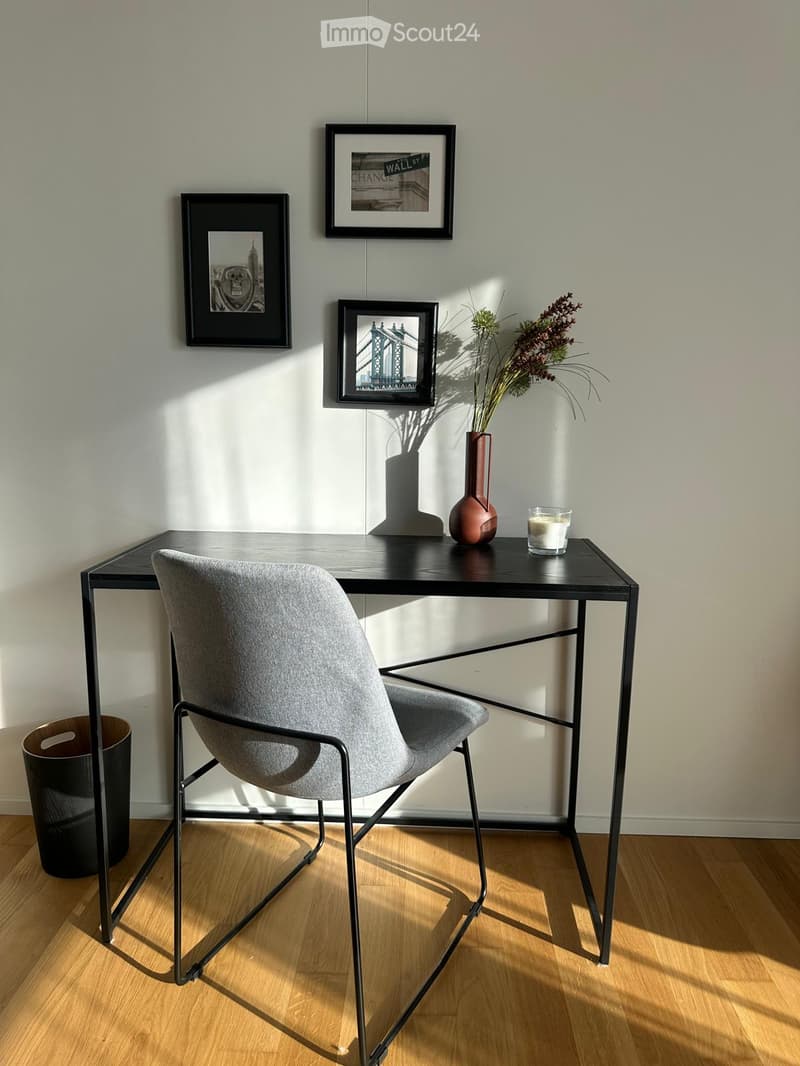 Modern, minimalist room with a grey upholstered chair, black metal-framed table, vase with dried flowers, candle, and three picture frames on the wall.