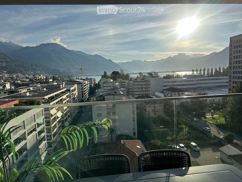 Balcony with a view of a cityscape, mountains, and a lake, featuring a table and chairs.