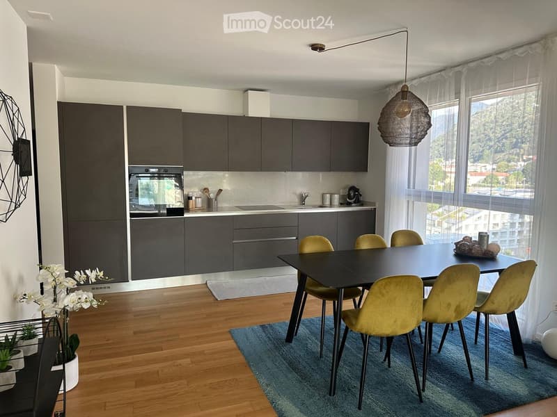 Modern kitchen with dark grey cabinetry, stainless steel oven, sink, and faucet. A black dining table with six yellow chairs sits on a blue rug. A pendant light hangs above the table, and large windows with white curtains offer a scenic view.