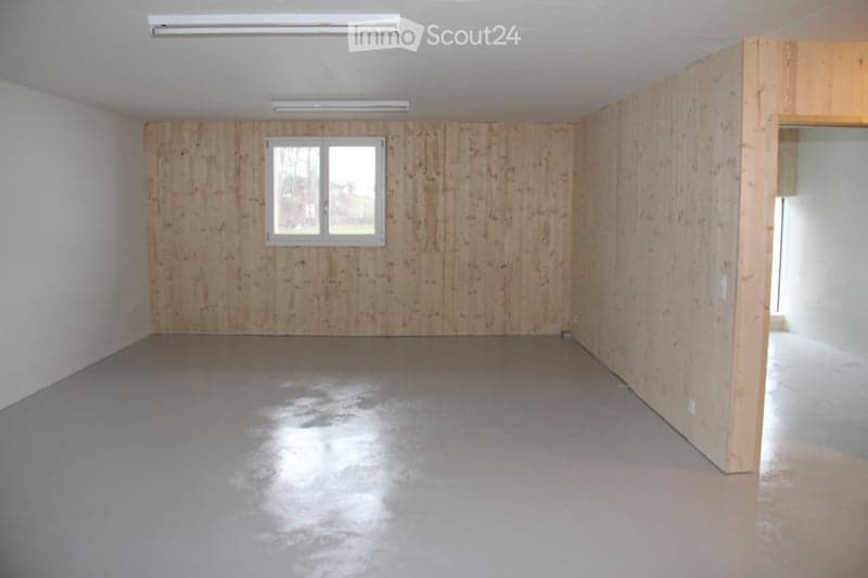 Empty room with wooden panel walls, grey concrete floor, and a single window. No furniture or equipment present.
