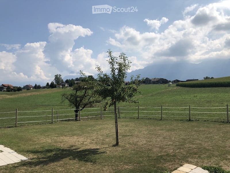 Open green field with a wooden fence, a few trees, and distant houses under a partly cloudy sky.